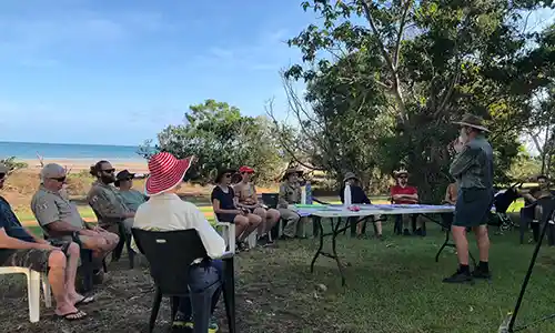 A small group sits in a semi‑circle on a grassy area near the beach while a presenter speaks under the shade of trees.