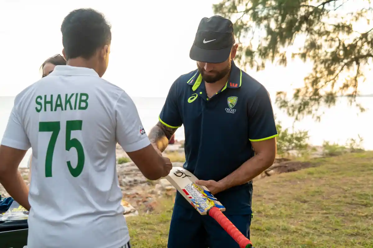 ricket player signs a bat for fan in Shakib 75 shirt by water at sunset.